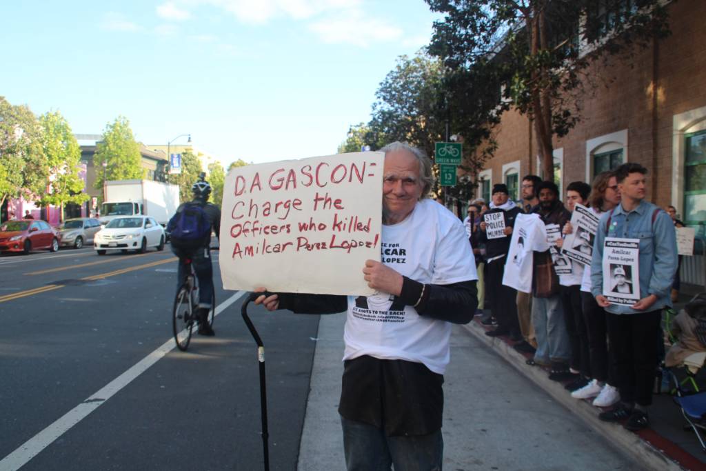 An elderly supporter stands with a placard demanding charges against the Police Officers involved in shooting of Amilcar Perez Lopez. Photo By - Sana Saleem