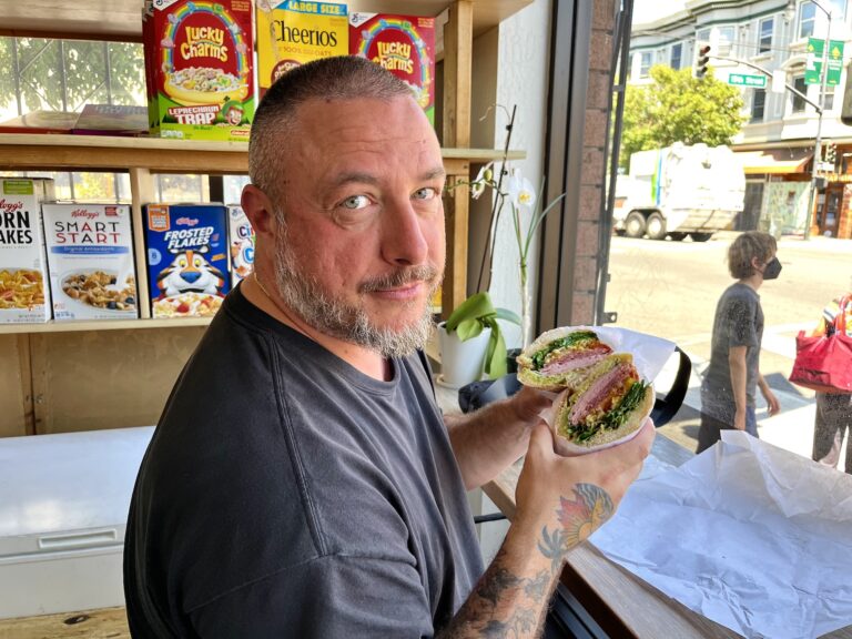 A bearded man holds up half a sandwich at a window counter, cereal boxes on the shelves behind him